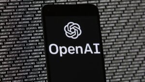 The OpenAI logo appears on a mobile phone in front of a computer screen with random binary data.