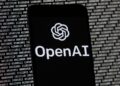 The OpenAI logo appears on a mobile phone in front of a computer screen with random binary data, March 9, 2023, in Boston.