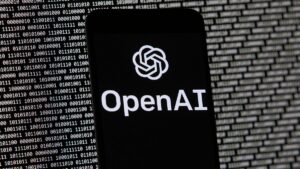 The OpenAI logo appears on a mobile phone in front of a computer screen with random binary data, March 9, 2023, in Boston.