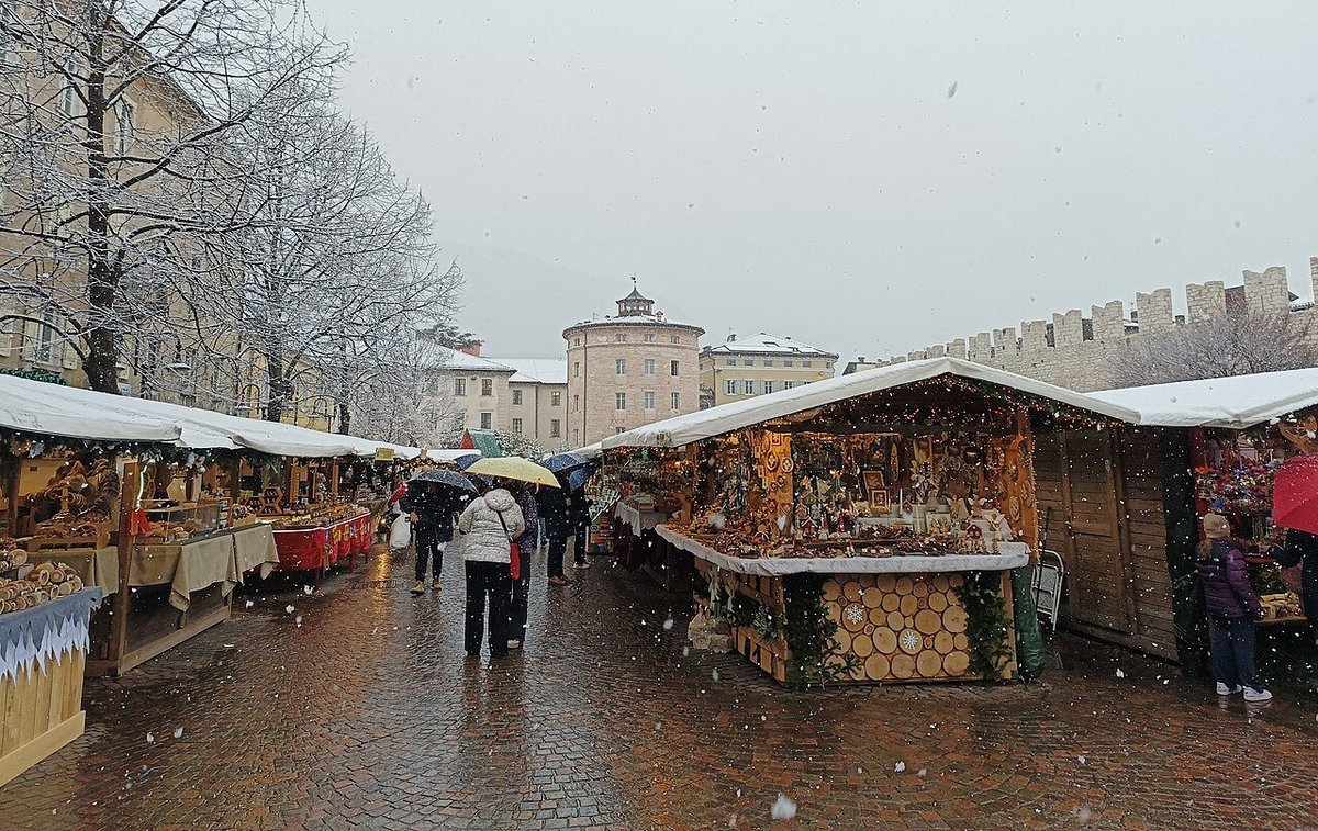 Casette di legno del mercatino di Natale di Trento in piazza Fiera sotto la neve, con le mura medievali sullo sfondo